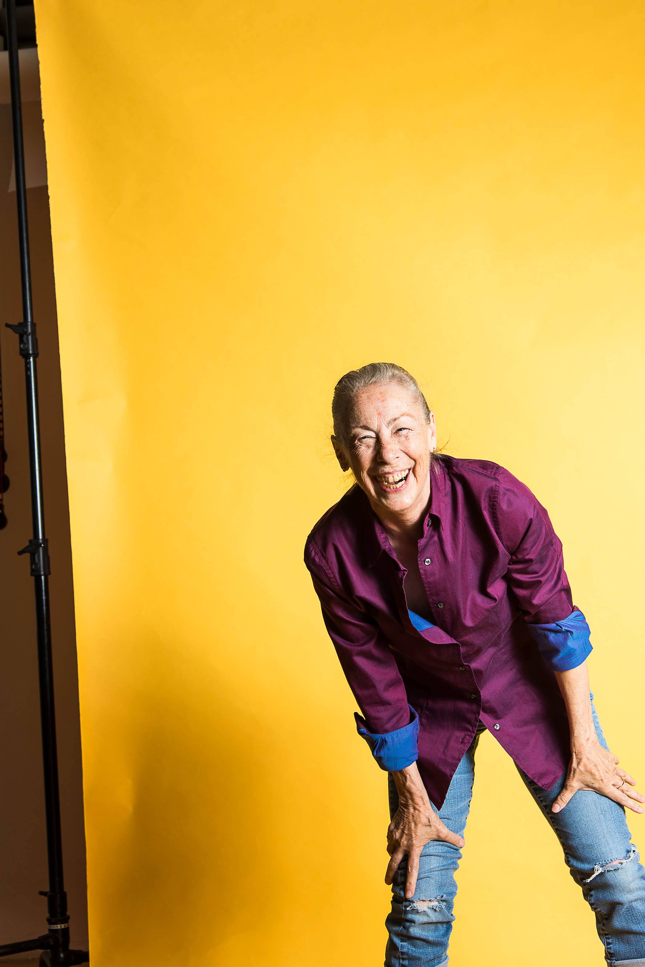 Debbie Wilson laughing, wearing a plum shirt and jeans against a bright yellow backdrop.